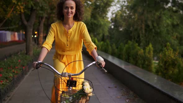 Beautiful Woman in Long Yellow Dress Riding a City Bicycle with a Basket and Flowers in the City