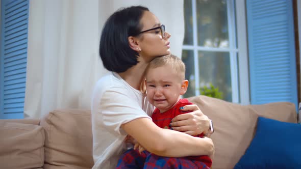 Young Mother Holding and Comforting Crying Little Son Sitting on Sofa at Home