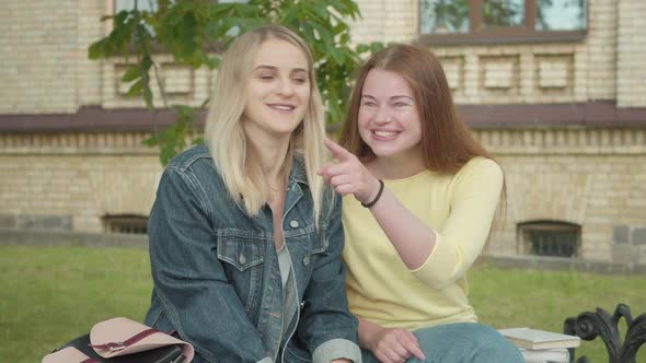 Portrait of Beautiful Joyful Female Students Pointing Away and Laughing. Front View of Two Young