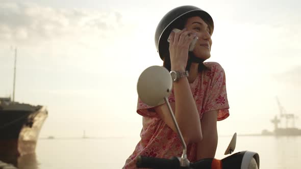 Young Beautiful Girl Speaking on Phone Near Sea on Scooter at Sunrise