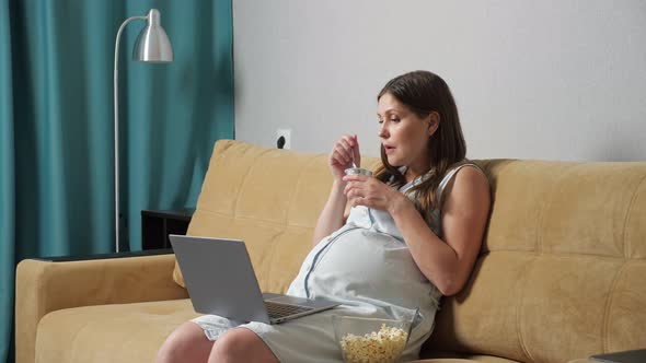 Pregnant Woman Eating Yogurt and Popcorn and Looking at Laptop While Sitting on Sofa