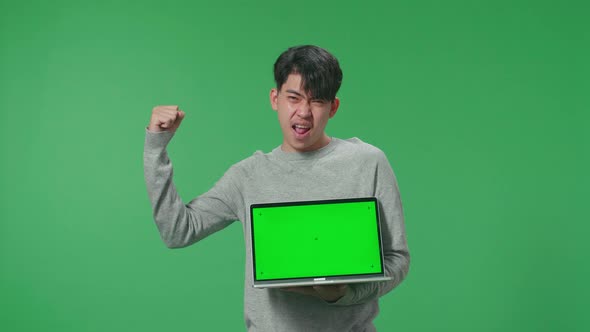 A Happy Asian Man Celebrating While Holding Green Screen Computer In The Green Screen Studio