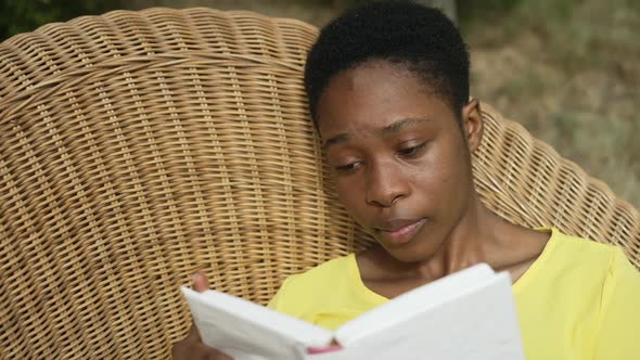 Closeup Absorbed African American Beautiful Woman Reading Book Lying on Wicker Sofa in Garden