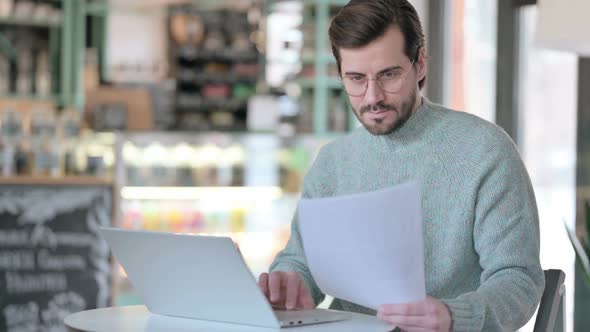 Young Man Reading Paper While Using Laptop Cafe