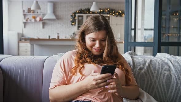 Relaxed Young Plus Size Woman Using Smartphone Surfing Social Media Sitting on Sofa at Home