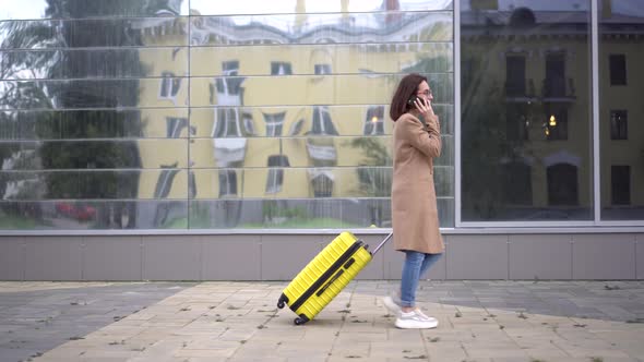 A Young Woman Walks with a Yellow Suitcase and Speaks on the Phone. A Girl with Glasses and a Coat