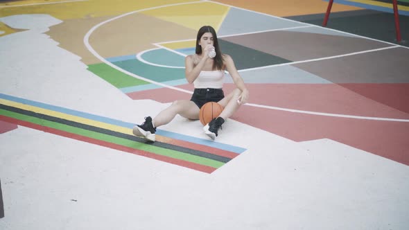 Extreme Wide Shot of Slim Young Sportswoman Sitting on Outdoor Basketball Court Drinking Refreshing