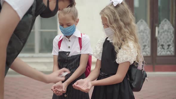 Schoolboy in Covid-19 Face Mask Spraying Hand Sanitizer on Palms of Schoolgirls. Caucasian