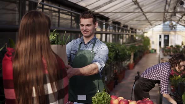 Portrait of European Salesman Wearing Green Apron Is Giving Organic Food in Brown Paper Bag To