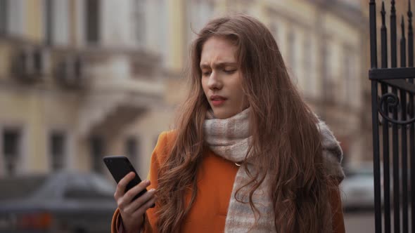 Portrait of Pretty Student Girl Wearing Coat and Scarf Holding Smartphone and Choosing Necessary
