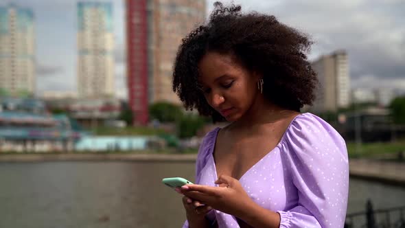 a Young African Woman Is Dressed in a Lilac Top with Ties and Puffy Sleeves. She Stands on the City