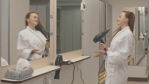 Young Charming Sportswoman in White Bathrobe Drying Hair with Dryer in Gym Locker Room. Confident