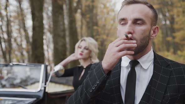Serious Portrait of Gentleman Stands in Front of His Lady and Smokes Cigar
