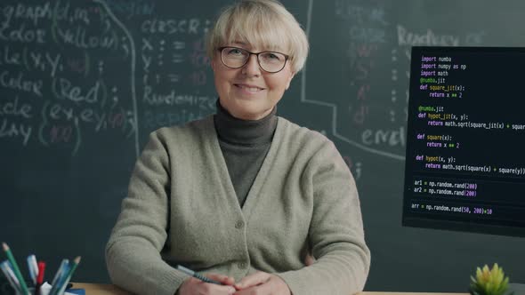 Portrait of Cheerful IT Teacher Sitting in Classroom with PC and Blackboard and Looking at Camera alt