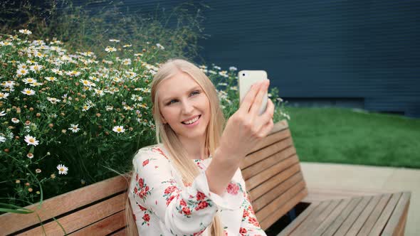 Lovely Young Female Using Smartphone To Make Video Call While Sitting on Bench on Living Roof