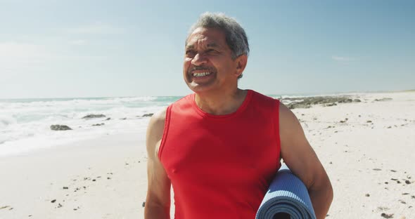 Portrait of hispanic senior man standing on beach, holding yoga mat and smiling