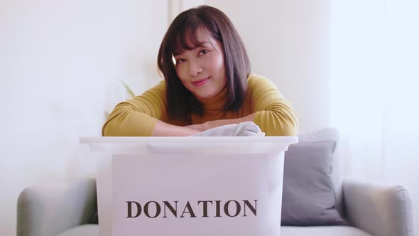 Portrait of young Asian woman looking at camera holding box with clothes for donation. Concept campa