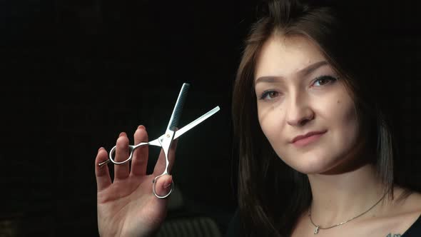 Happy Young Brunette Hairdresser Holding Scissors While Standing in Front of Camera Against Black