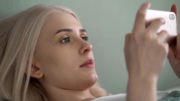 A Young Beautiful Woman Lies on a Bed with a Phone in Her Hands and Looks at the Screen