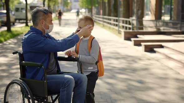 Close Up of the Disabled Father in Mask Sitting on the Wheel Chair Wearing Mask to His Son Before