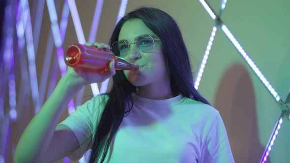 Middle Shot of Young Carefree Woman Drinking Colorful Alcohol and Smiling at Camera in Night Club