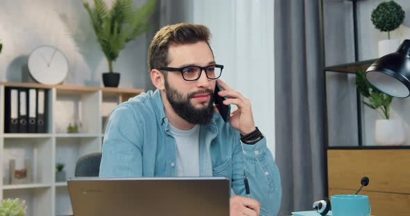 Guy in Glasses Talking on Mobile Sitting in front of Computer in His Home Office