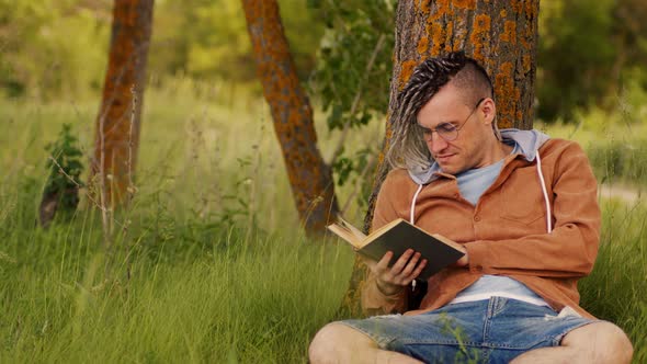 A Man in Casual Clothes and Glasses Reads Literature Under a Tree