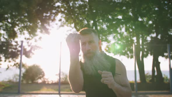 Middle Aged Man with Long Gray Beard Boxing with Shadow on Basketball Court During Sunrise Slow