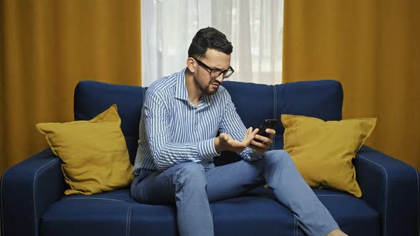 Displeased Man Looks Angrily at Smartphone Types Text Messages on Sofa