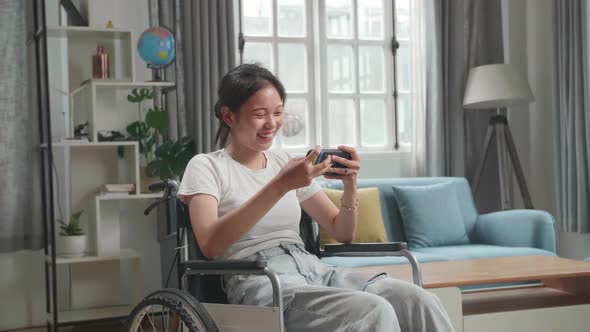 Asian Woman Sitting In A Wheelchair While Use Mobile Phone Playing Video Game At Home