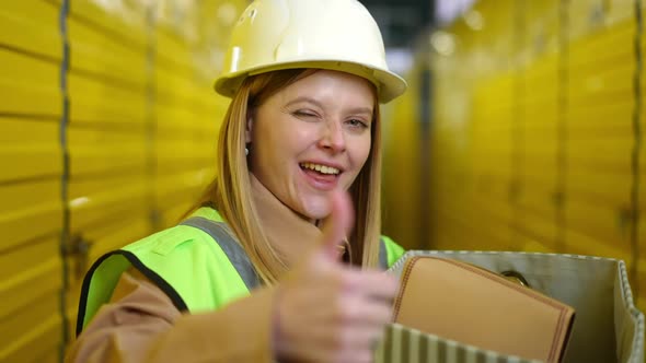 Satisfied Beautiful Woman Winking Gesturing Thumb Up Standing with Box in Yellow Storage