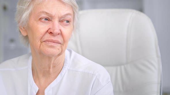 Old wise grey haired mother with blue eyes looks aside with sad face