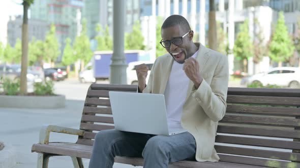 African Man Celebrating Success on Laptop While Sitting Outdoor on Bench