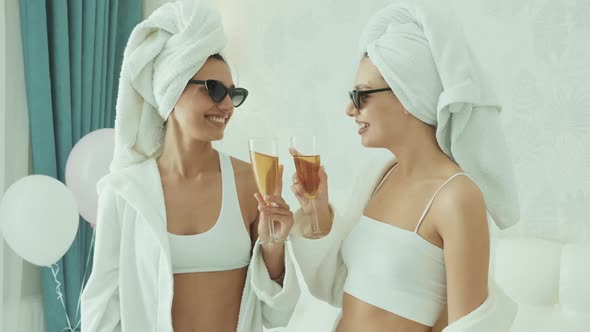 Two young beautiful smiling women in white bathrobes and towels on head having party indoors