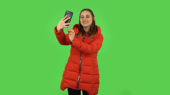 Lovely Girl in a Red Down Jacket Is Making Selfie on Mobile Phone Then Looking Photos. Green Screen