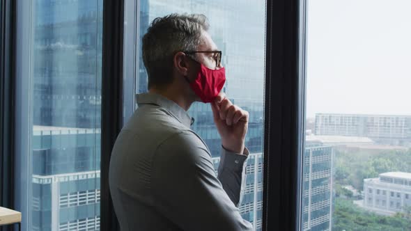 Thoughtful caucasian man wearing face mask looking out of window at modern office