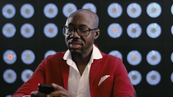 Smiling Bald Africanamerican Businessman in Red Suit Looks at Smartphone