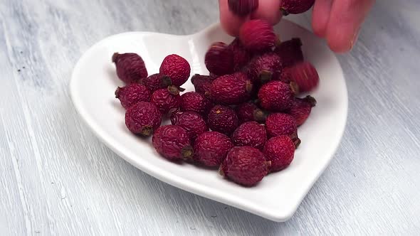 Hands pour dried red rosehips into a white plate with a heart shape in slow motion.