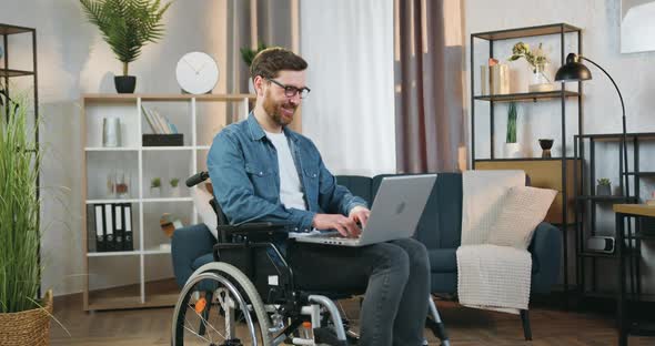 Man in Glasses Sitting in Wheelchair After Accident at Home and Working on Computer