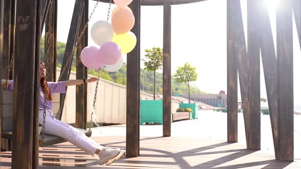 Happy Red-haired Girl Having Fun Carefree Sitting and Swaying on Swing with Fountain of Balloons.