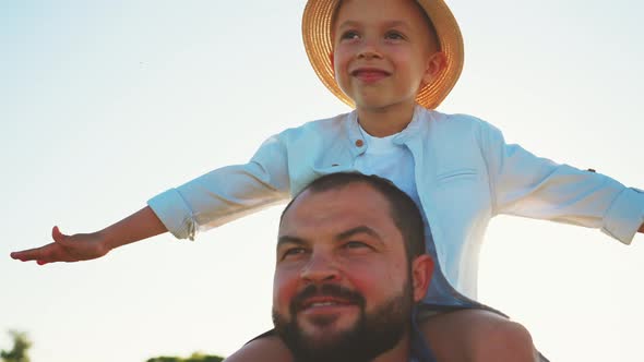 Close Up Front View Father Carrying Son on Shoulders in Field Boy Rejoices and Spreads Arms to Sides