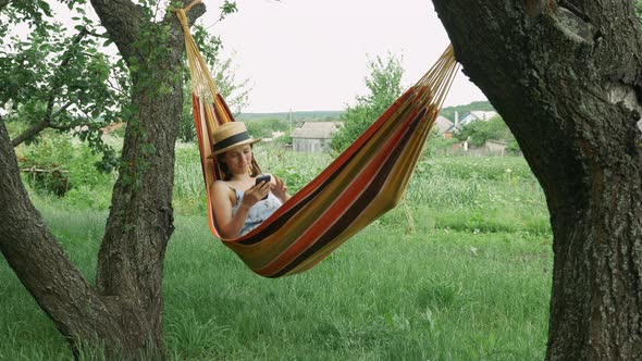 Relaxed young woman looking at mobile phone in hammock outdoors. Girl in hat and dress with phone