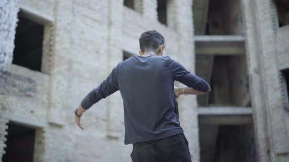 Back View of Aggressive Young Man Drinking Beer and Shouting at Abandoned Slum. Mixed-race Alcoholic