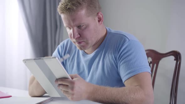 Portrait of Thoughtful Caucasian Man Looking at Tablet Screen and Writing Down Notes, Smart Young