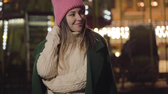 Smiling Caucasian Girl in Pink Hat and White Sweater Standing Outdoors and Looking Around. Beautiful