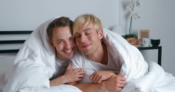 Young Gay Couple Lying on Bed Together in Bedroom Hugging and Kissing