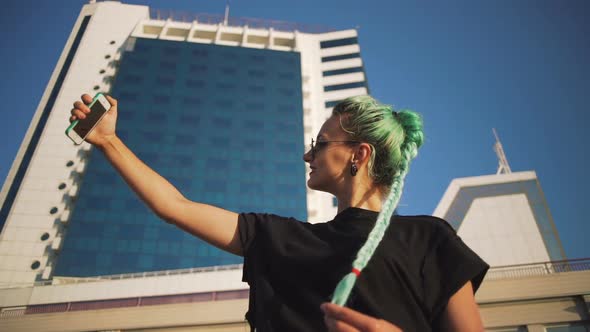 Portrait of Young Stylish Hipster Woman with Green Hair Using Smartphone on Urban Buildings