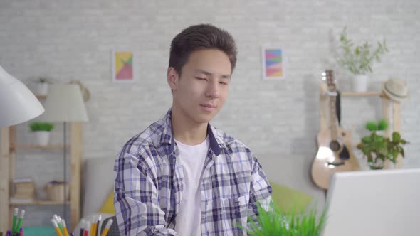 Young Asian Man Working at Laptop in Living Room at Home and Looking at Camera