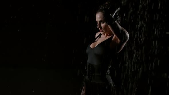 A Young Busty Woman in a Black Sexy Suit Poses Beautifully in a Dark Studio and in the Rain