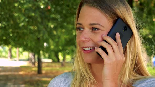 Young Pretty Blond Woman Phones with Smartphone - Park with Trees in Background - Closeup